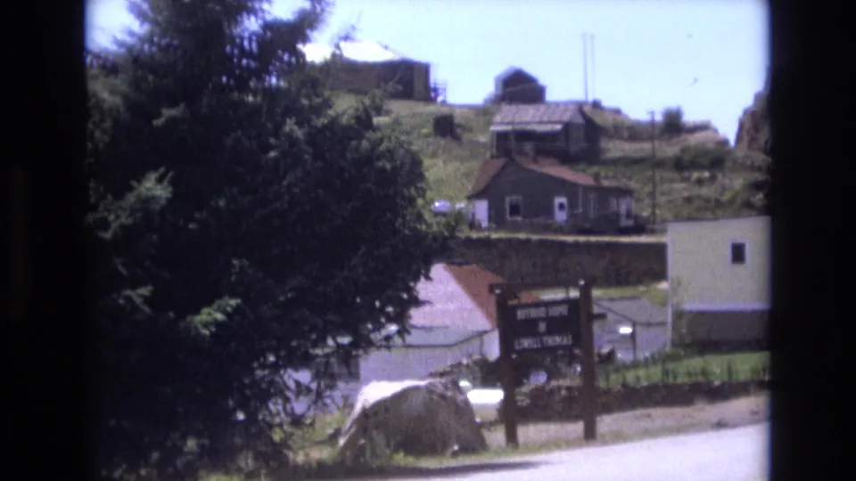 1973 Colorado Springs 8mm frame showing a roadside sign, trees, a light-colored building, and small structures higher on the hill.
