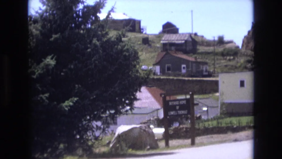 1973 Colorado Springs roadside frame with an identifiable sign, a pale building, trees, parked cars, and small hillside structures above.