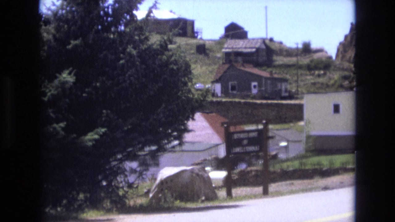1973 Colorado Springs home-movie frame showing a large evergreen, a paved road, hillside houses, a stone wall, and a roadside wooden sign.