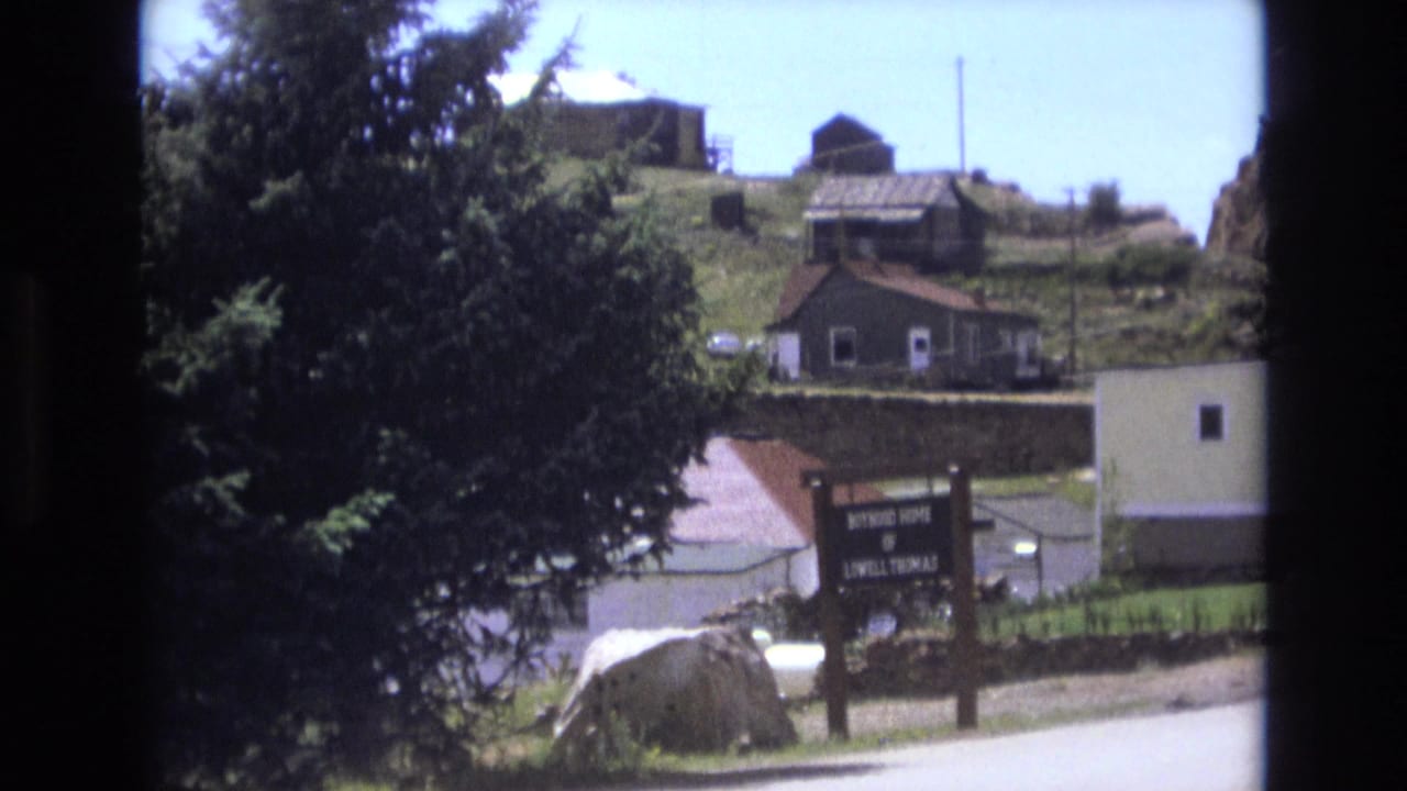 Roadside wooden sign below hillside houses, partly framed by a large evergreen tree.