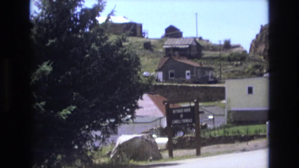 1973 Colorado Springs 8mm frame showing a tree-dominated roadside view, wooden sign, stone wall, and small residential buildings on a grassy incline.