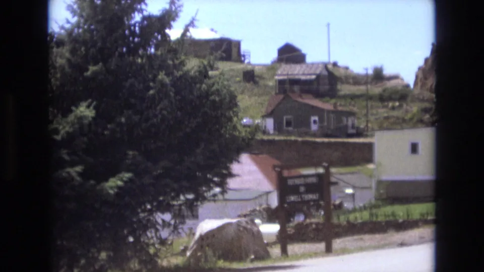 1973 Colorado Springs home-movie frame with a large evergreen in the foreground, a wooden roadside sign, and modest houses set into a grassy hillside.