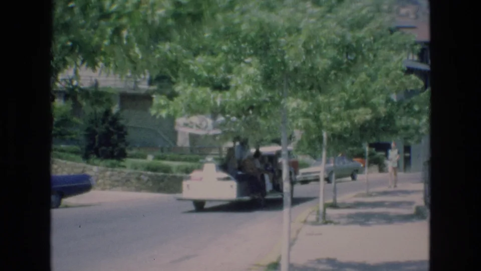 A handheld 1973 Colorado Springs zoo view shows a leafy walkway or drive with parked cars, distant visitors, stone edging, and buildings partly obscured by trees.