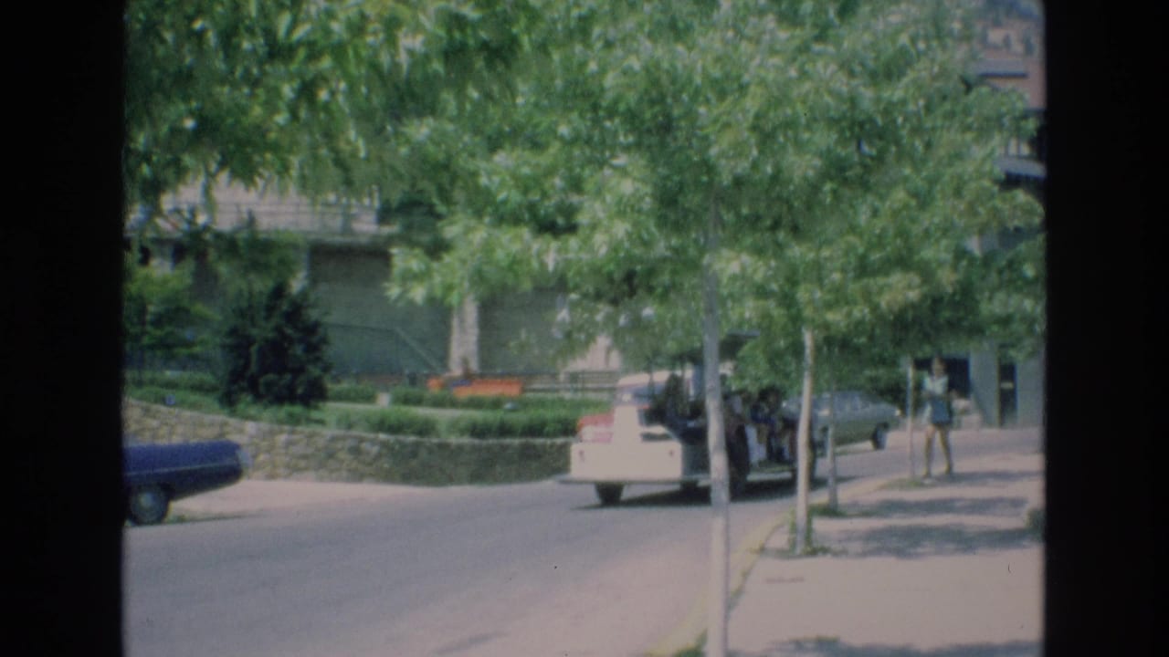 In Colorado Springs, Colorado, in 1973, a white open-air tour vehicle travels down a leafy street with a blue parked car and a pedestrian on the sidewalk.