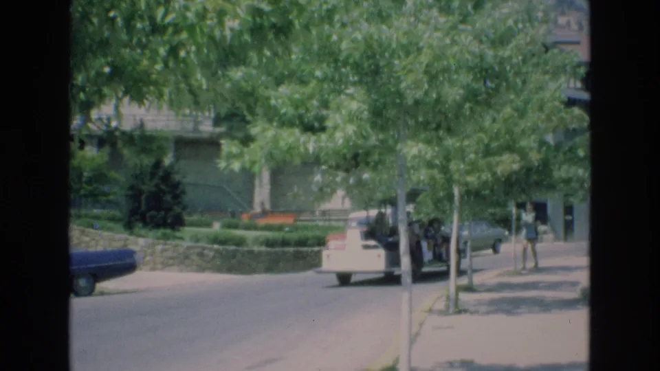 In Colorado Springs, Colorado, in 1973, a white open-air tour vehicle travels down a leafy street with a blue parked car and a pedestrian on the sidewalk.