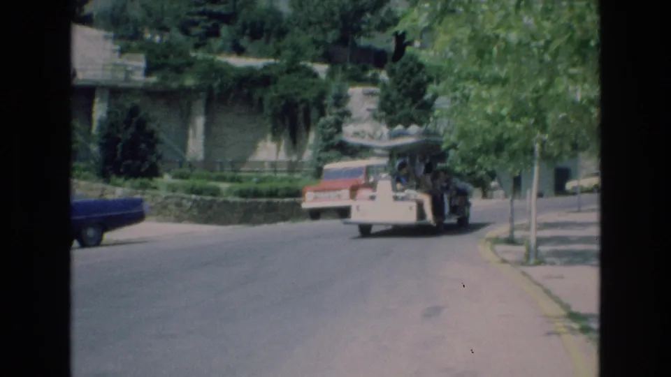 A rear view captures the open-air tour vehicle driving away on a quiet Colorado Springs street in 1973, framed by trees, a stone wall, and a parked blue car.