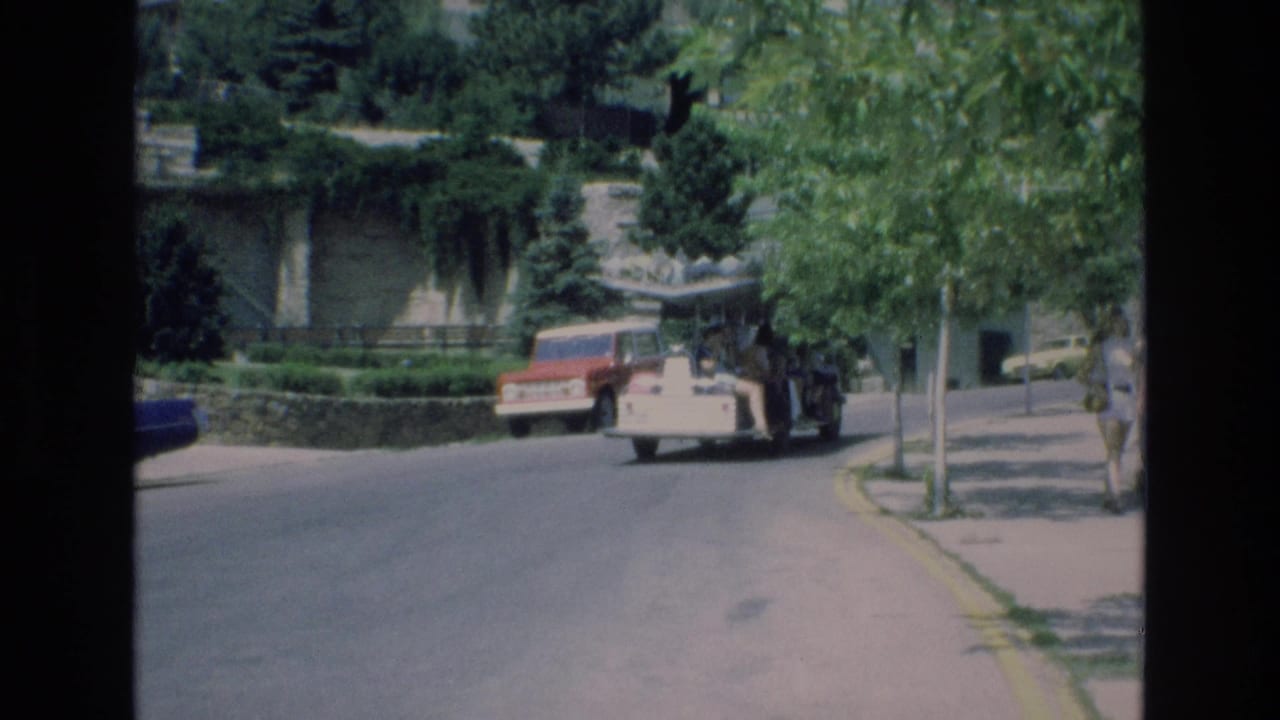 1973 Colorado Springs zoo footage shows a white open-air people mover traveling on a paved road past trees, walls, and a parked red vehicle.