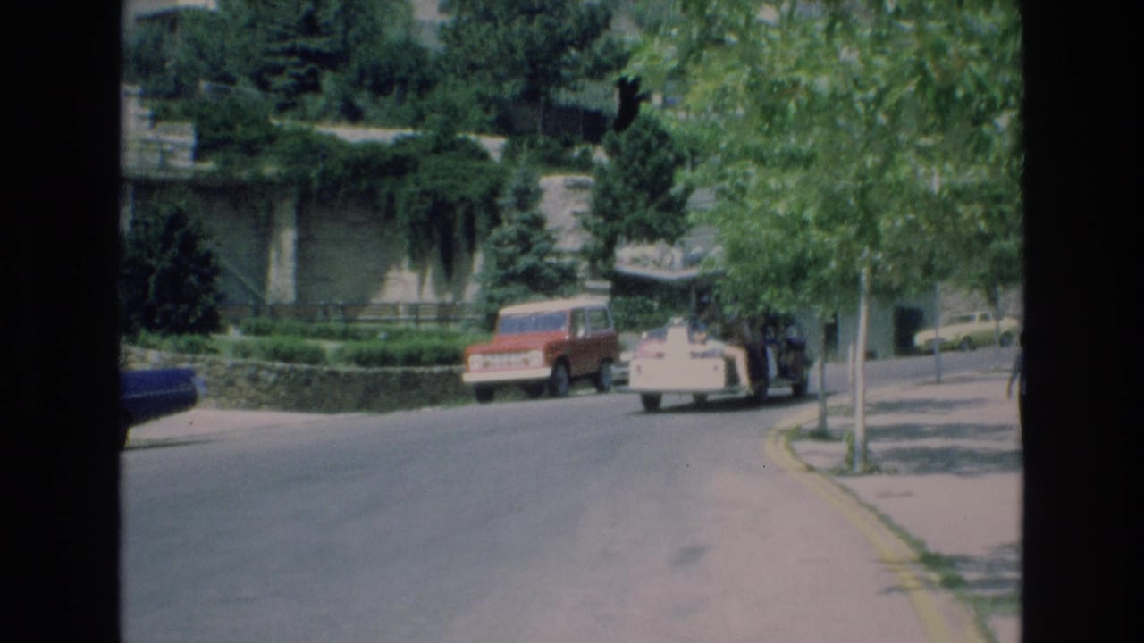 People mover continues along the road beside a parked red vehicle and trees.