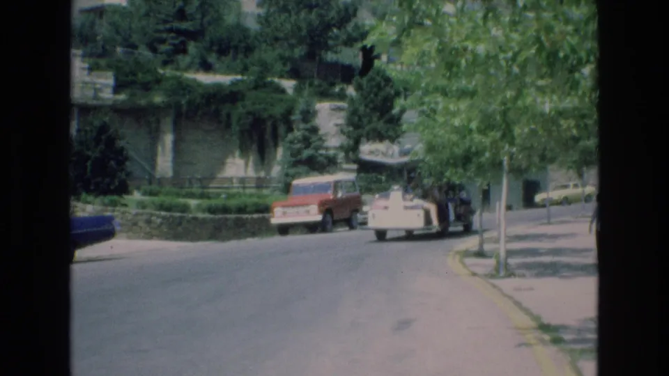 The white zoo people mover appears farther along the paved road in Colorado Springs, with a red vehicle parked nearby and trees shading the scene.