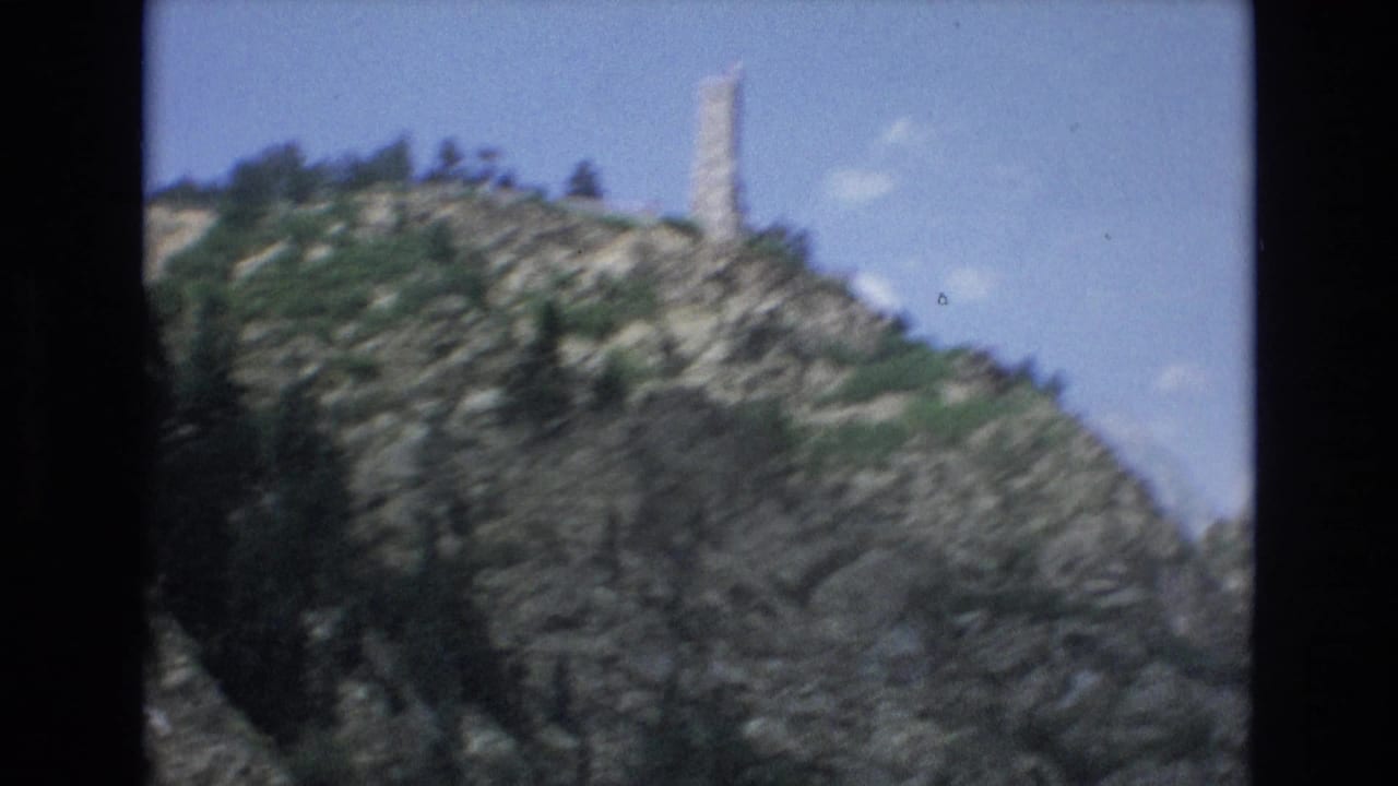 1973 Colorado Springs home-movie frame showing a pale stone tower rising above a rocky hillside with evergreen trees and blue sky.