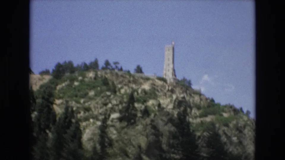 1973 Colorado Springs frame showing the tower at the right of a rocky, green hillside with trees along the crest and a clear blue sky behind it.