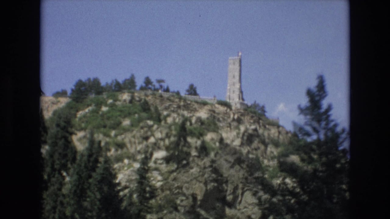 1973 Colorado Springs 8mm frame showing a tall stone tower on a rocky, tree-lined hill beneath a clear blue sky.