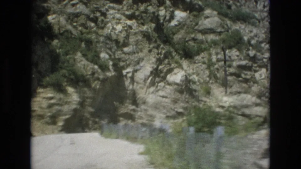 Colorado Springs 8mm frame showing the lower rocky hillside, evergreen trees, brush, and a pale road or path at the bottom of the shot.