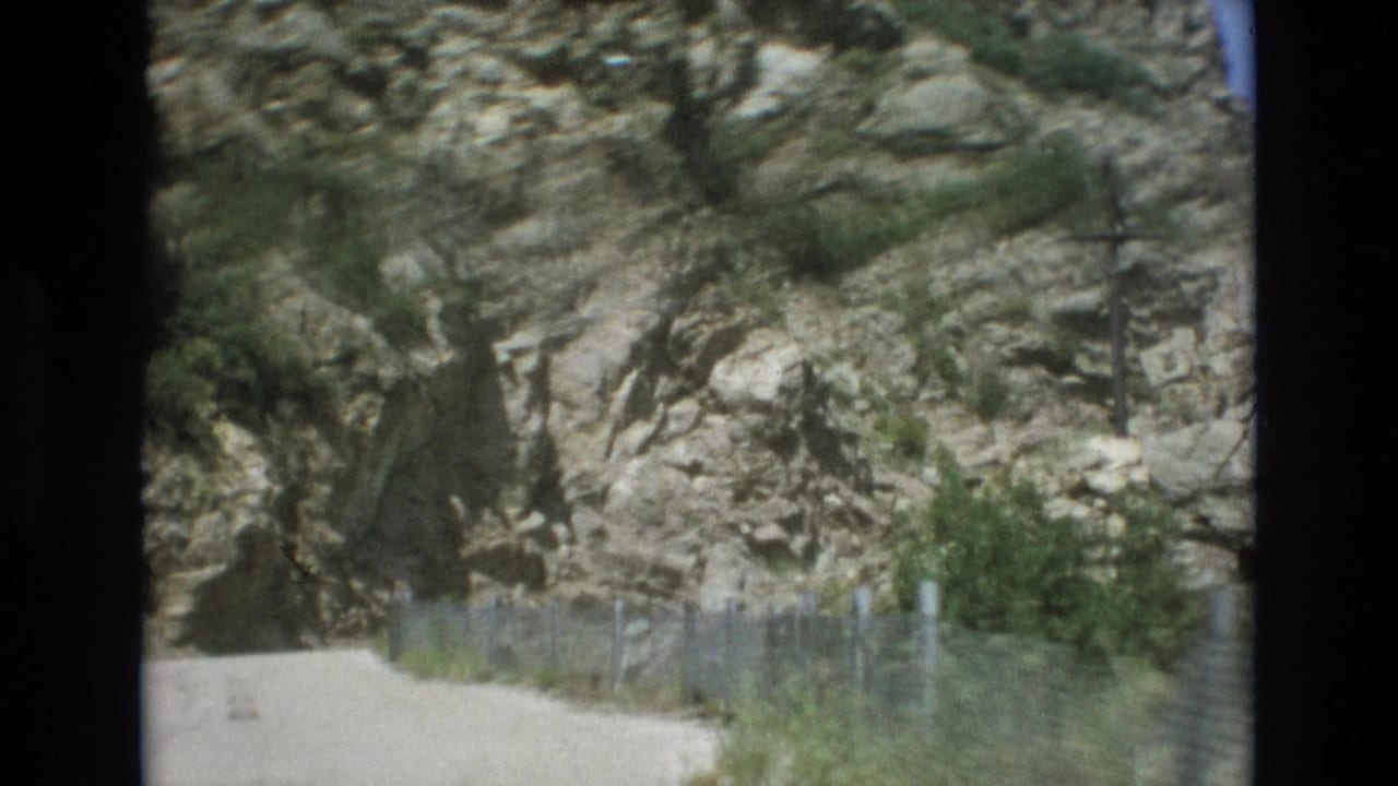 1973 Colorado Springs home-movie frame showing a pale dirt path curving along a rugged rock face with wire fencing and scattered shrubs.