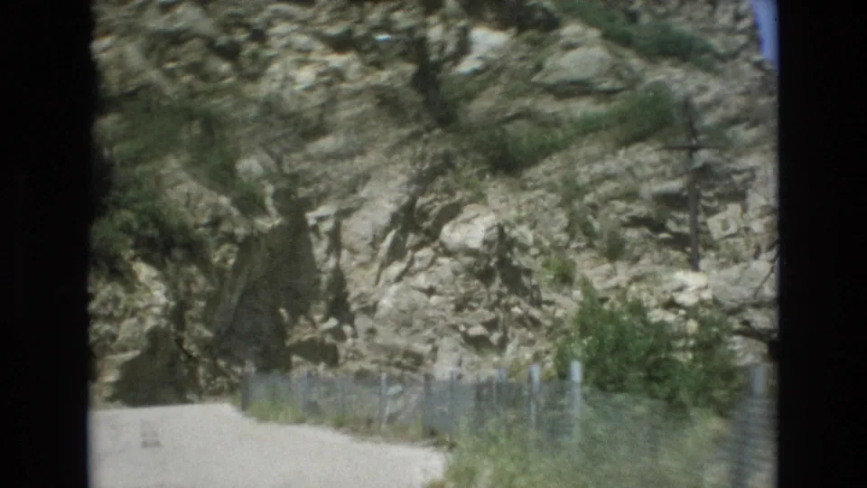 1973 Colorado Springs home-movie frame showing a pale dirt path curving along a rugged rock face with wire fencing and scattered shrubs.