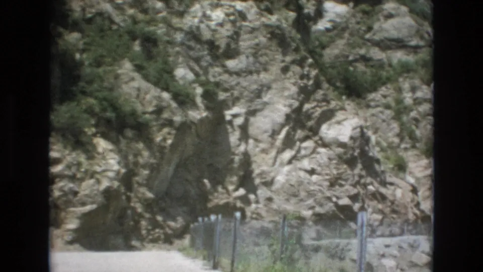 1973 Colorado Springs home-movie frame showing the rocky slope closer in, with the dirt path and wire fence continuing along the bottom of the view.