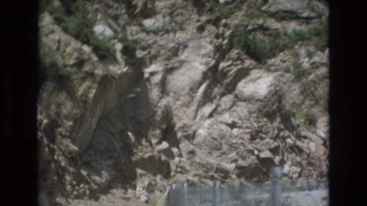 1973 Colorado Springs-area travel frame showing a pale rocky cliff face above a faint chain-link roadside guard fence.