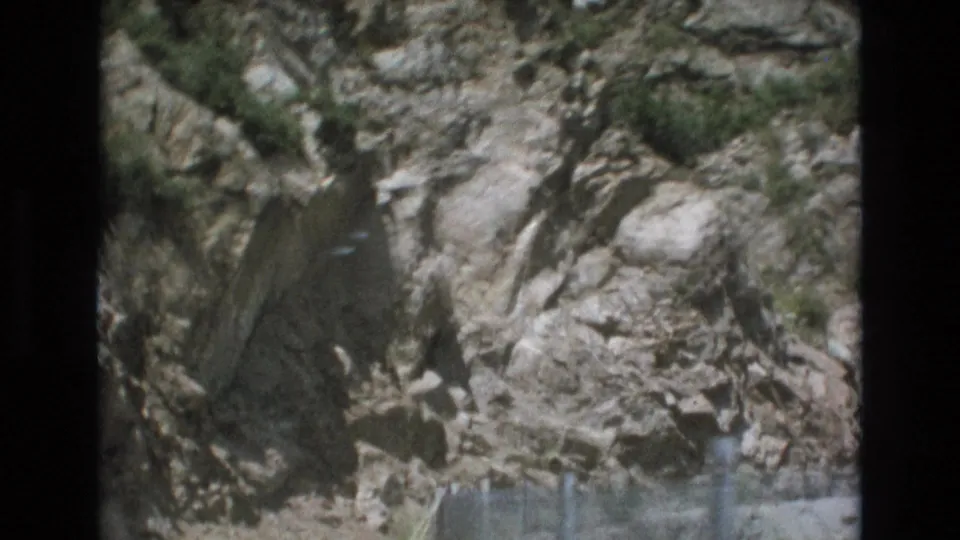 1973 Colorado Springs-area travel frame showing a pale rocky cliff face above a faint chain-link roadside guard fence.