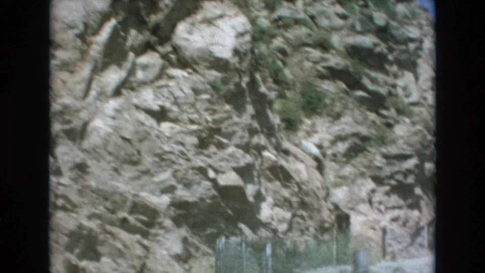 Soft 8mm frame of a pale rocky slope, scattered greenery, and roadside fencing during a Colorado Springs-area drive.