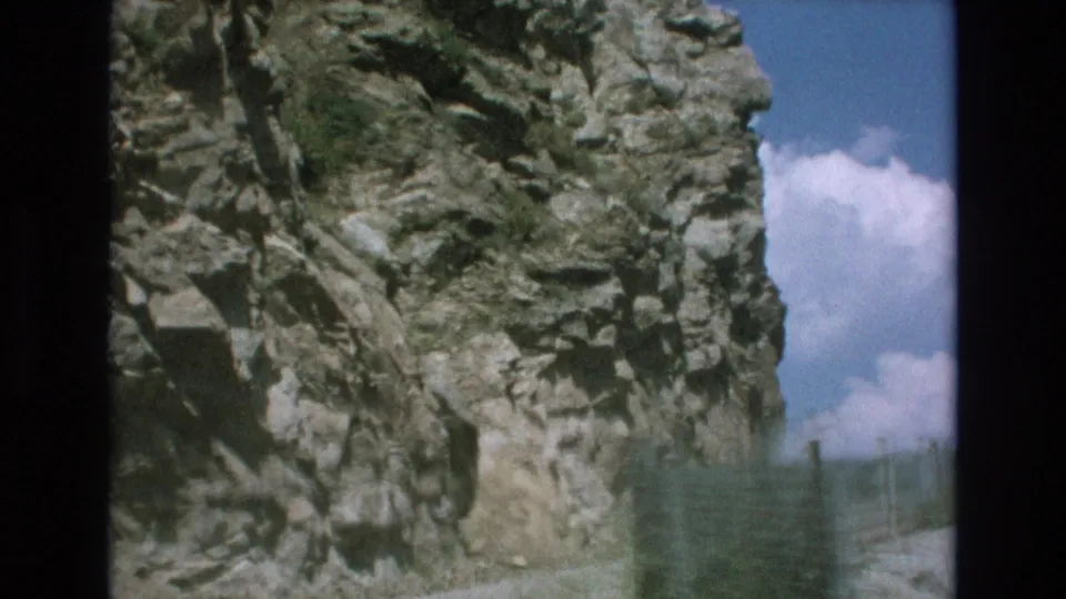Final frame showing a cliffside rock face, roadside guard fence, and blue sky with clouds beyond the steep edge.