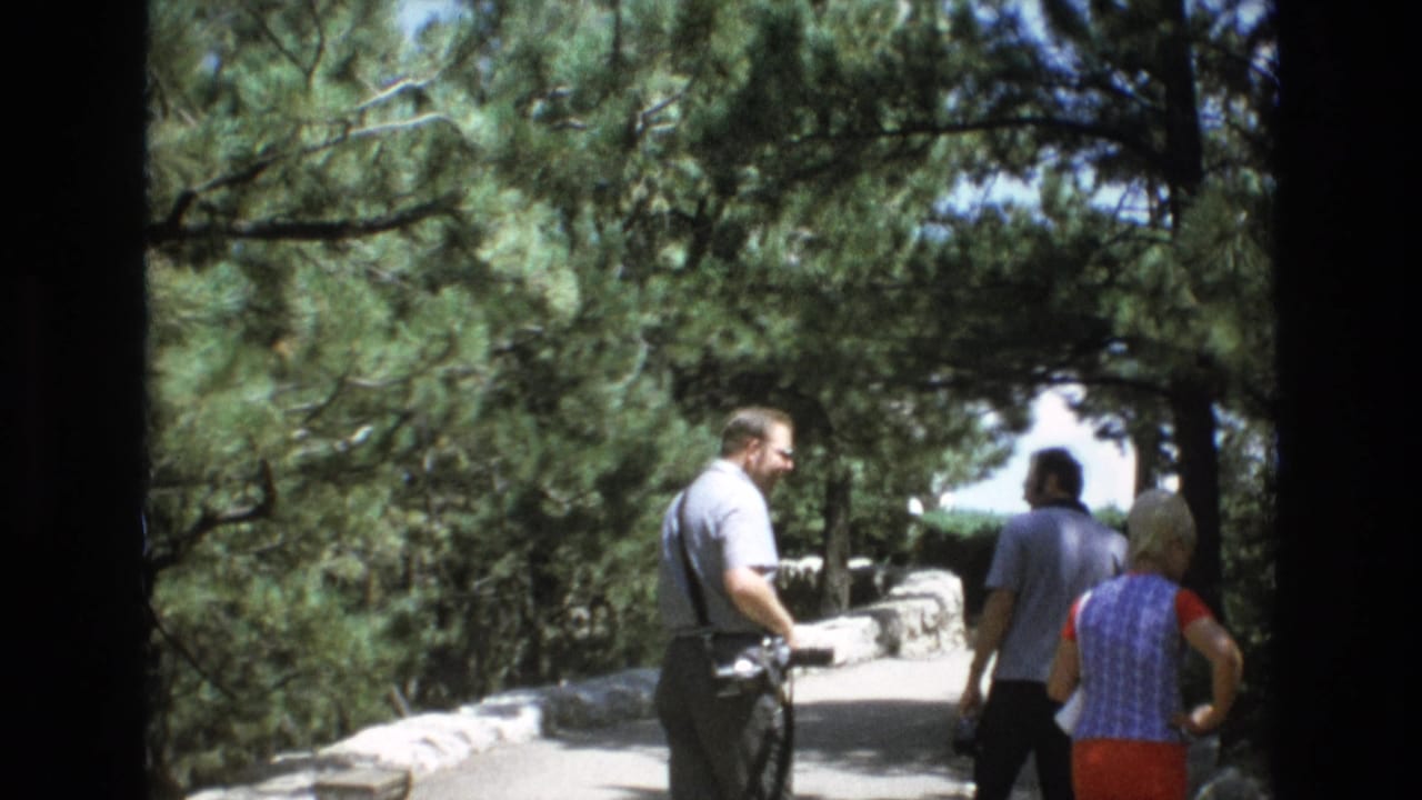 In Colorado Springs according to metadata, adult tourists walk along a stone-lined path beneath pine trees during a sunny 1970s sightseeing visit.