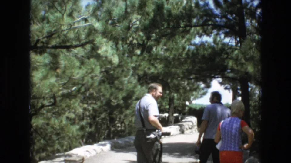 In Colorado Springs according to metadata, adult tourists walk along a stone-lined path beneath pine trees during a sunny 1970s sightseeing visit.