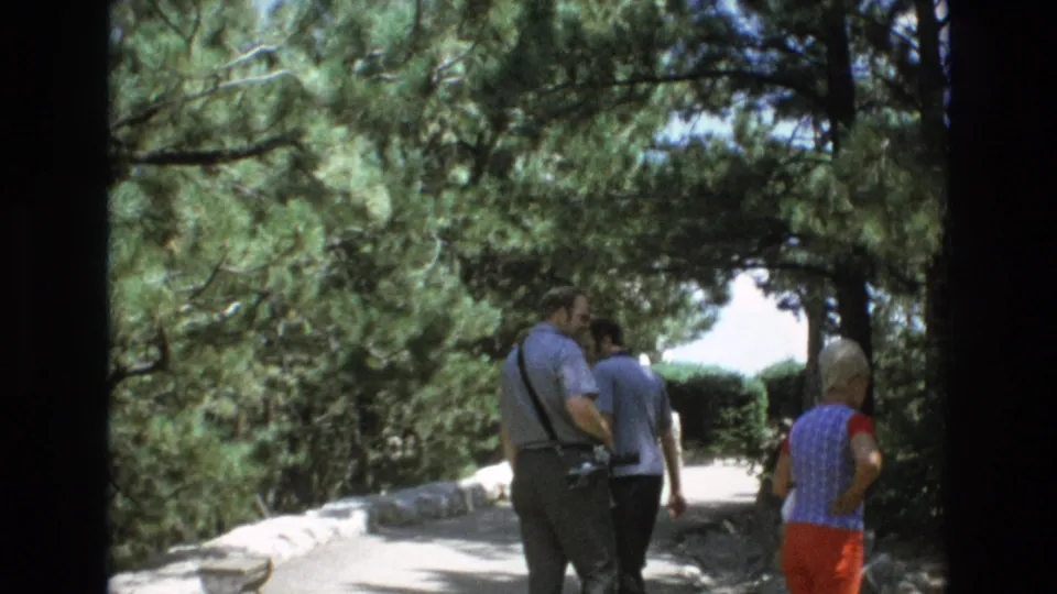 Adult visitors, including a woman in red pants and two men in casual shirts, move away along a shaded Colorado Springs park path.