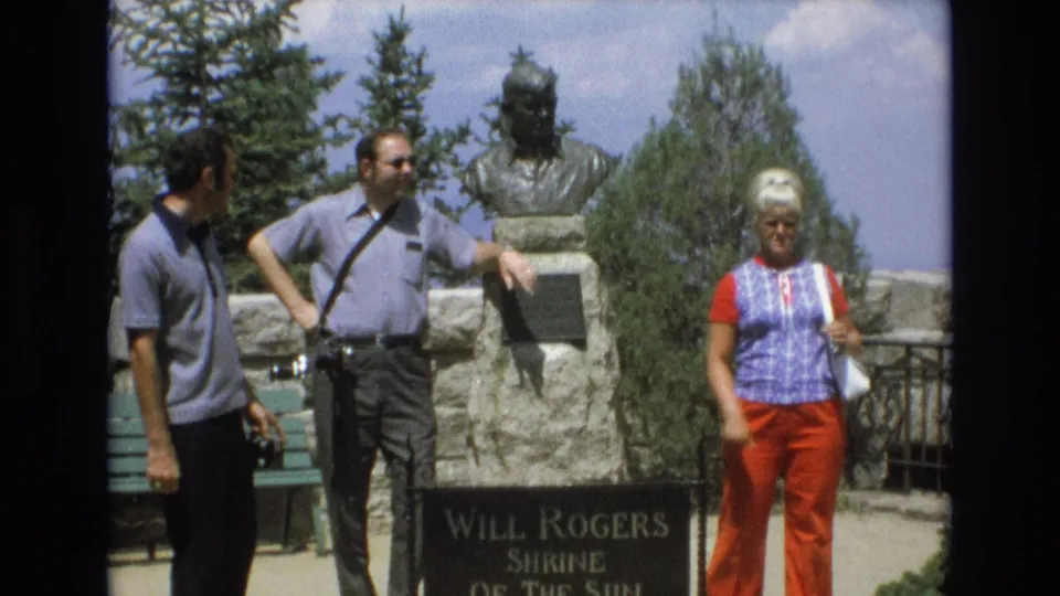 A man with a camera, another visitor, and a woman in red pants appear beside the Will Rogers Shrine of the Sun marker in bright outdoor light.