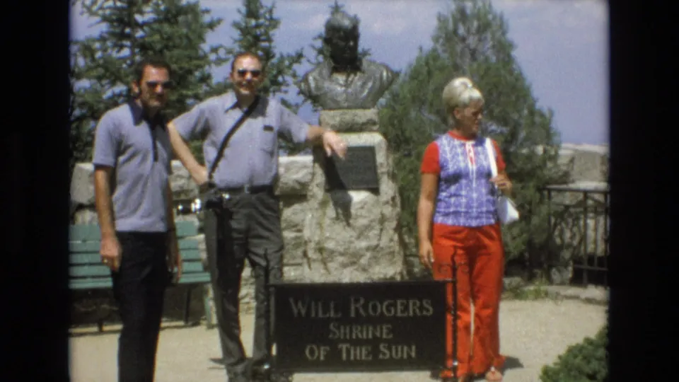 Two men wearing sunglasses and a woman in red pants pose beside the Will Rogers Shrine of the Sun sign and bust in Colorado Springs.