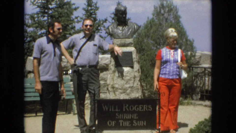 A 1973 home-movie frame shows three adult visitors at the Will Rogers Shrine of the Sun, with the woman turned slightly and the men standing near the stone monument.