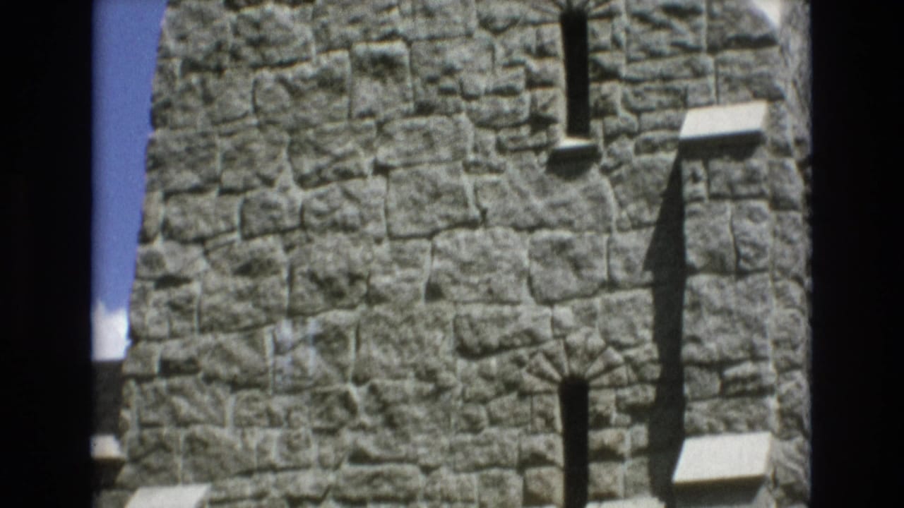 Upper stone tower wall with narrow slit windows and strong shadows against blue sky.