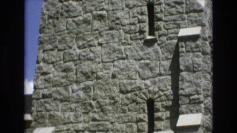 1973 Colorado Springs 8mm frame focused on the upper gray stone tower wall, with slim vertical openings, small ledges, and dark shadows cast across the masonry.