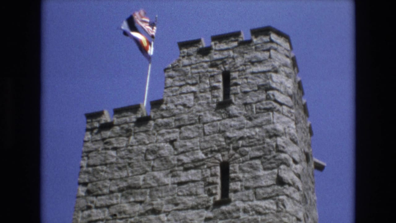 Stone tower top with a flagpole and flags against a clear sky.
