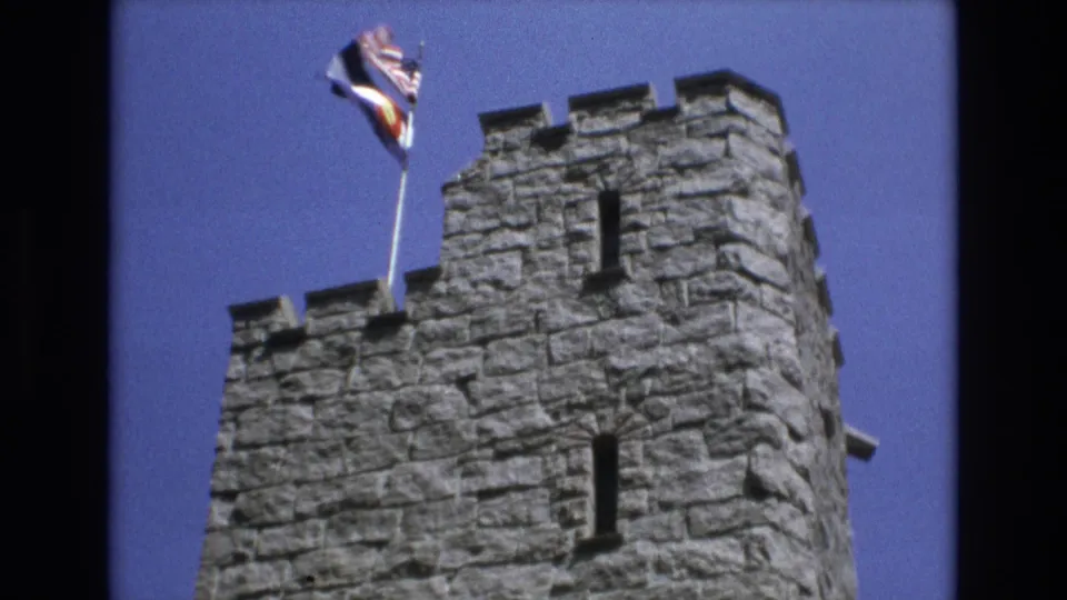 1973 Colorado Springs home-movie frame showing the upper part of a crenellated stone tower with flags waving from a pole above it.