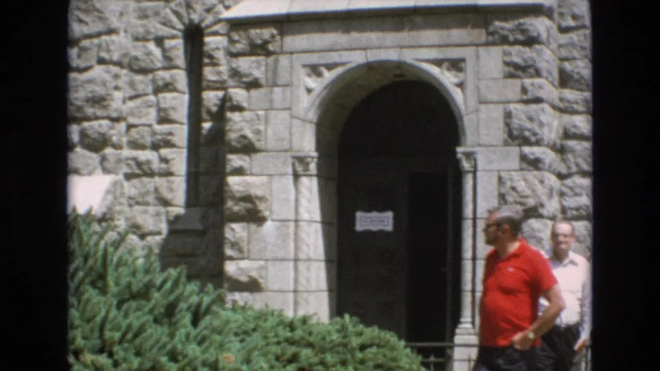 1973 Colorado Springs 8mm frame showing an arched stone doorway, green shrubs, narrow windows, and two adult men walking past the entrance.