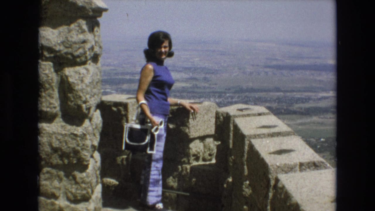 Adult woman in 1973 Colorado Springs stands beside a stone overlook wall, holding a white purse and facing the camera against a broad city and plains view.