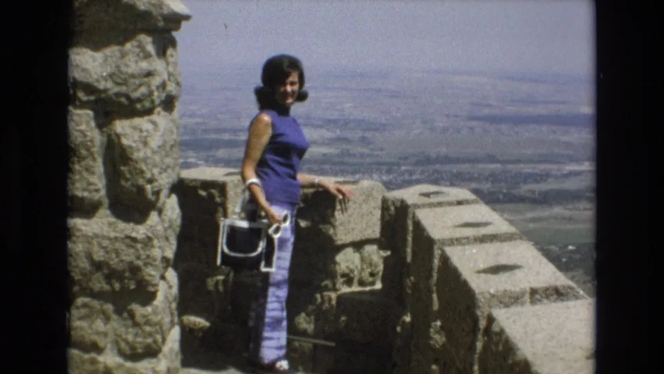 Adult woman in 1973 Colorado Springs stands beside a stone overlook wall, holding a white purse and facing the camera against a broad city and plains view.