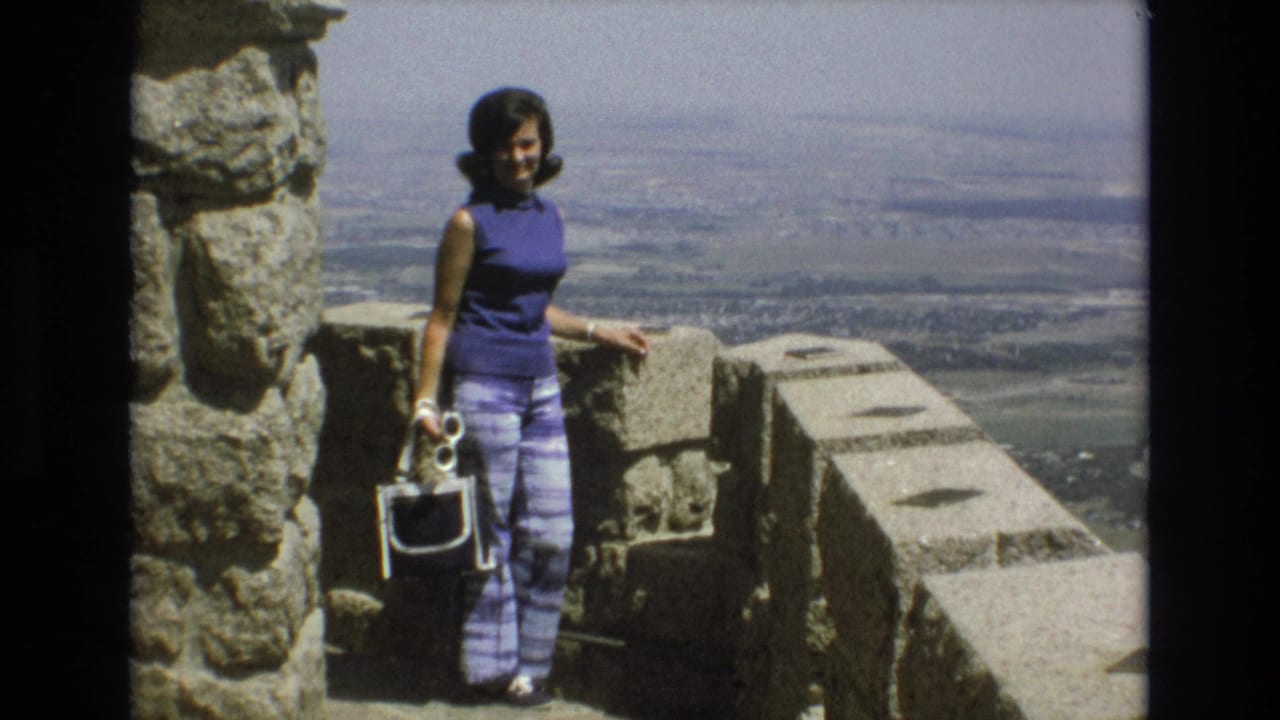 Woman poses near a stone wall and pillar with a panoramic view in the background.
