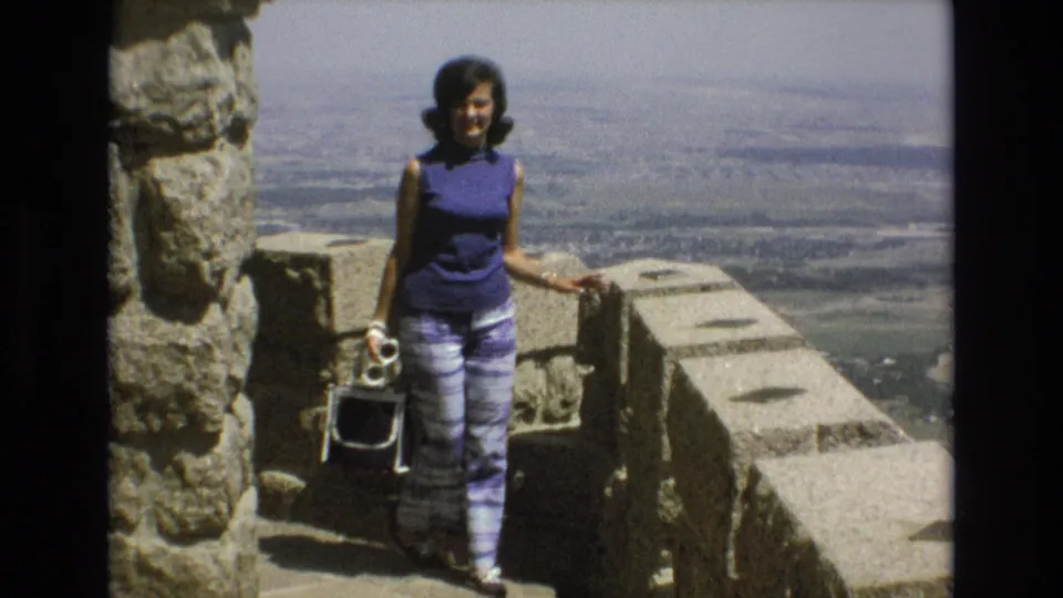 Adult woman at a 1973 Colorado Springs overlook stands near a rough stone pillar, with her white purse at her side and the sunlit landscape beyond the wall.