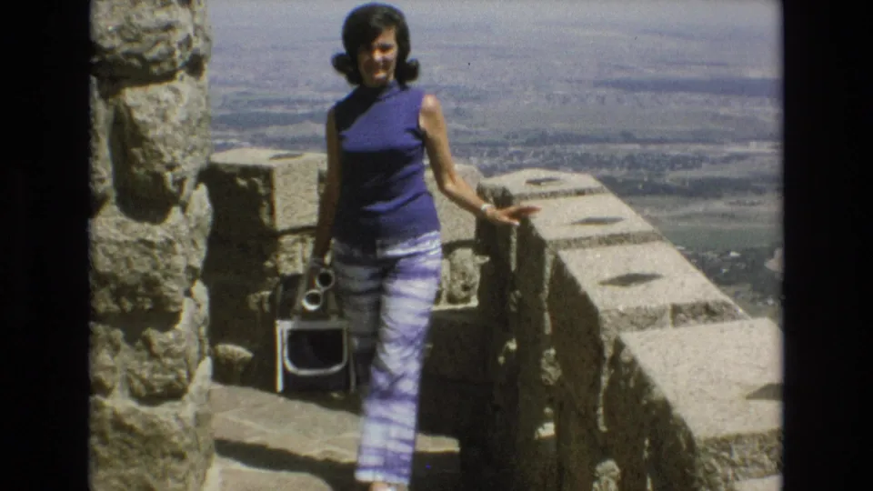Adult woman in 1970s clothing poses with one hand on a stone overlook wall in Colorado Springs, holding a white purse as the distant landscape fills the background.
