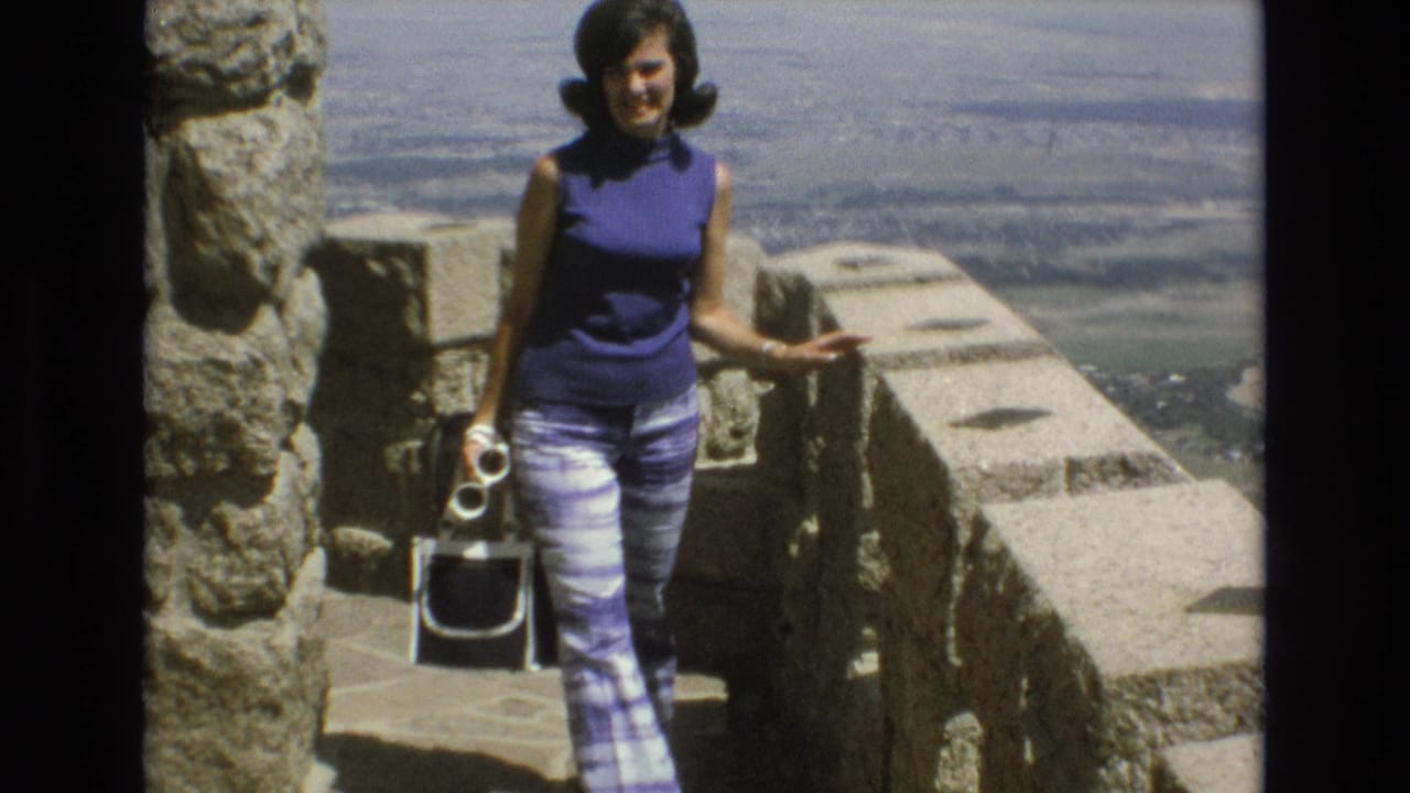 In supported 1973 Colorado Springs 8mm footage, a woman in a purple sleeveless top and patterned pants stands on stone steps at an overlook while holding a camera.