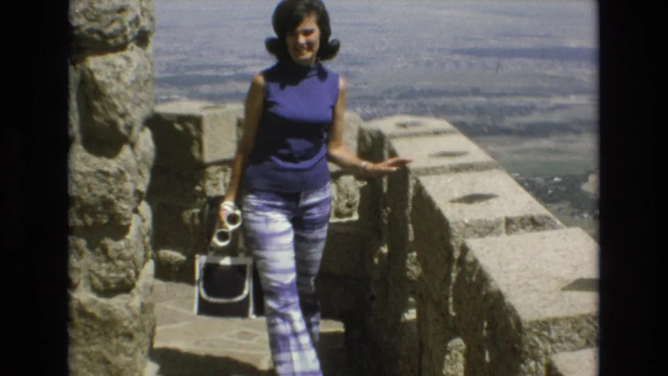 In supported 1973 Colorado Springs 8mm footage, a woman in a purple sleeveless top and patterned pants stands on stone steps at an overlook while holding a camera.