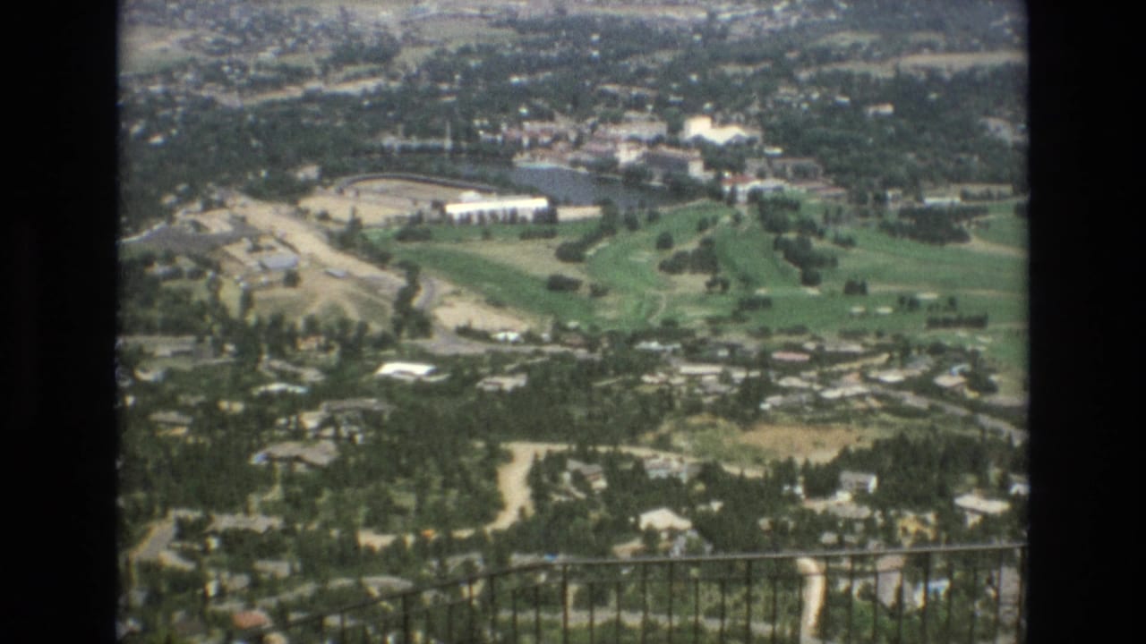 The camera looks down from an overlook toward Colorado Springs, showing roads, scattered buildings, green recreational land, and a foreground railing.