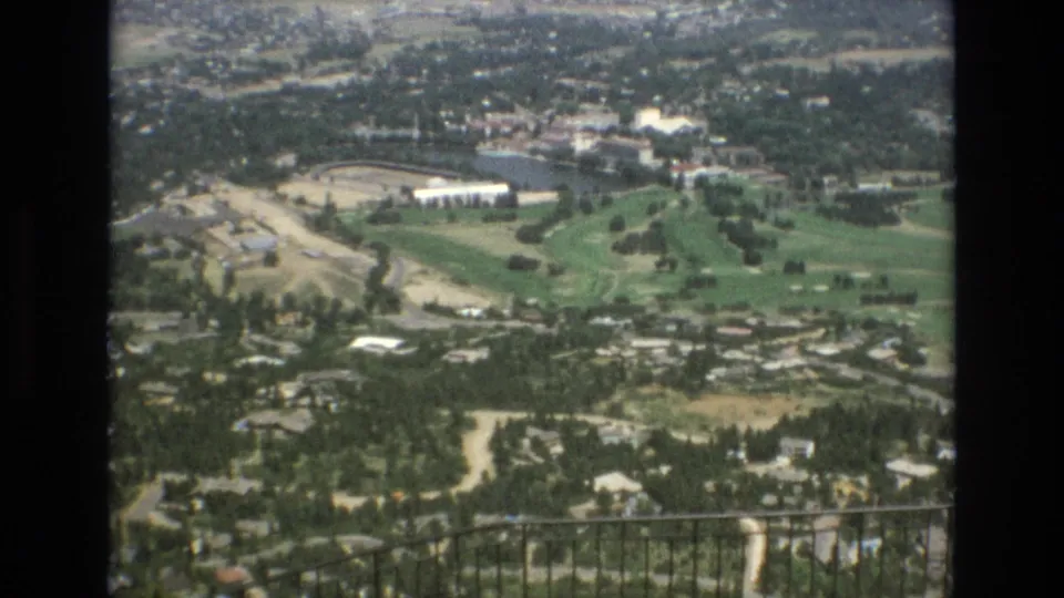 The camera looks down from an overlook toward Colorado Springs, showing roads, scattered buildings, green recreational land, and a foreground railing.
