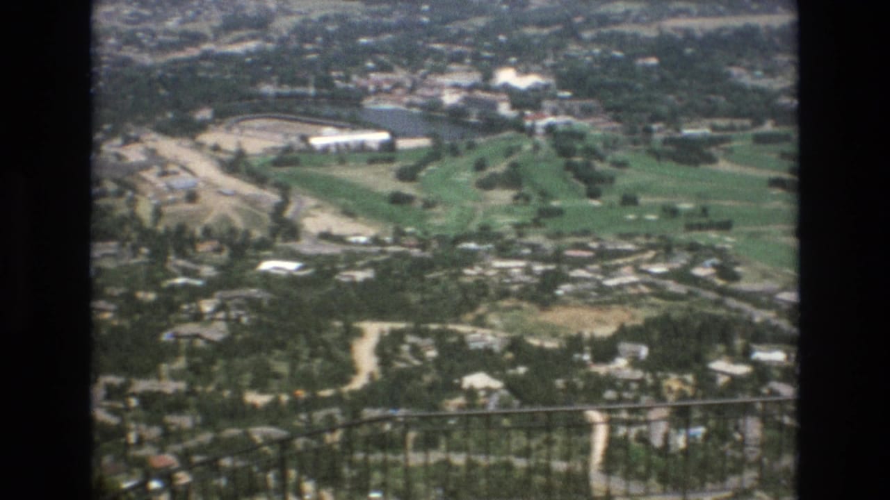 1973 Colorado Springs 8mm frame showing a high overlook across a green golf course, wooded residential area, pale roads, and a dark iron railing in the foreground.