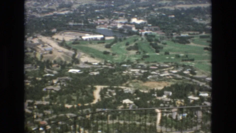 1973 Colorado Springs 8mm frame showing a high overlook across a green golf course, wooded residential area, pale roads, and a dark iron railing in the foreground.