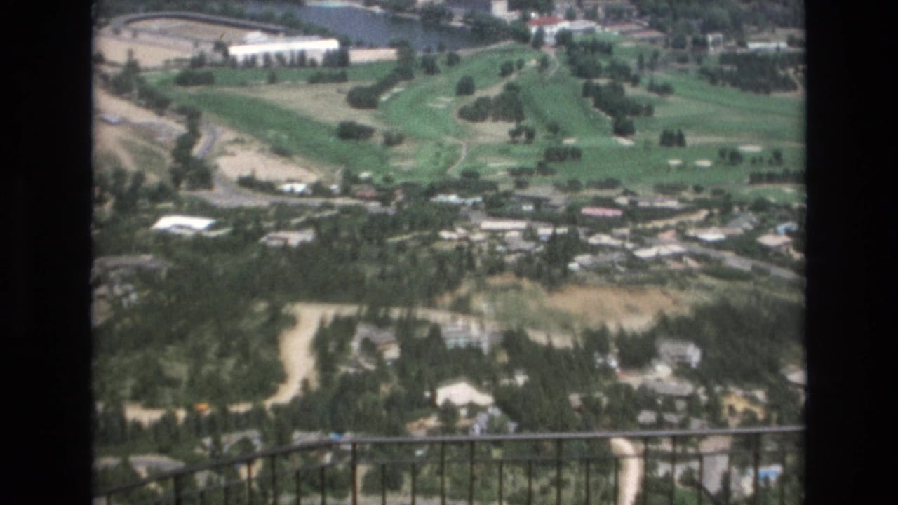 Elevated view of golf fairways, trees, roads, buildings, and an iron railing.