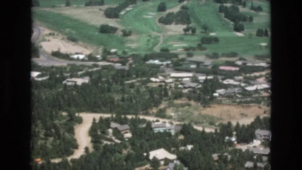 1973 Colorado Springs 8mm frame showing a closer high-angle view of a green golf course beside wooded homes, curving roads, and dry open ground.