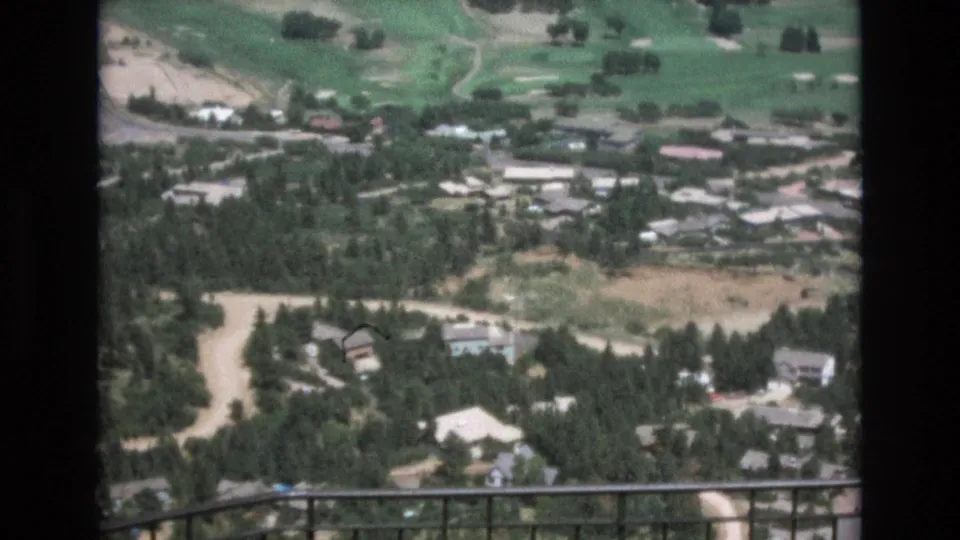 1973 Colorado Springs 8mm frame looking over wooded houses, winding roads, landscaped golf course edges, and an iron railing from an elevated viewpoint.