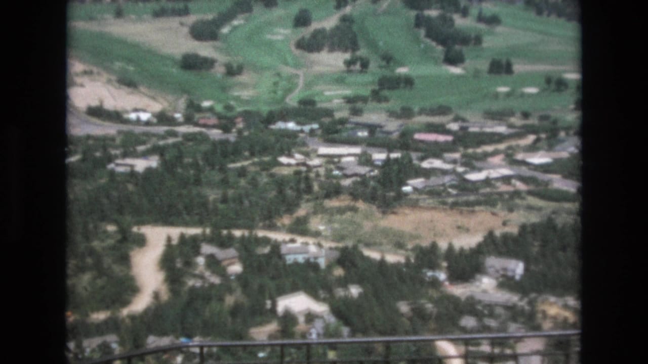1973 Colorado Springs home-movie frame showing a high vantage view over scattered homes, forested slopes, winding roads, and green golf-course fairways behind a foreground railing.